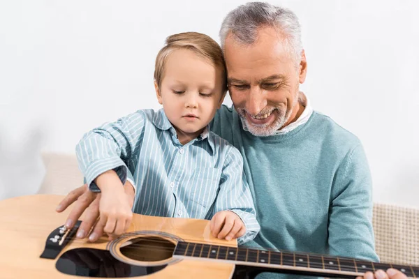 Красивый дедушка играет с внуком на коленях на акустической гитаре дома — стоковое фото