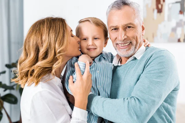 Selektiver Fokus eines fröhlichen Paares mittleren Alters, das seinen entzückenden Enkel hält und ihn auf die Wangen küsst — Stockfoto