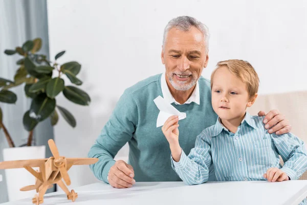 Hombre de mediana edad y nieto divirtiéndose con avión de papel en casa - foto de stock