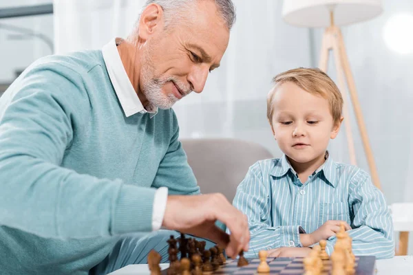 Селективное внимание мужчины средних лет, обучающего внука играть в шахматы дома — стоковое фото
