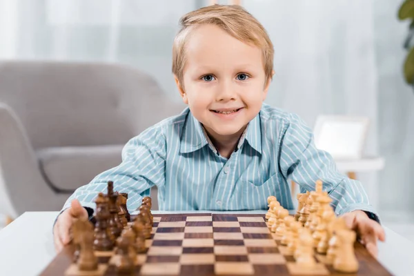 Retrato de niño feliz mirando la cámara cerca del tablero de ajedrez en casa - foto de stock