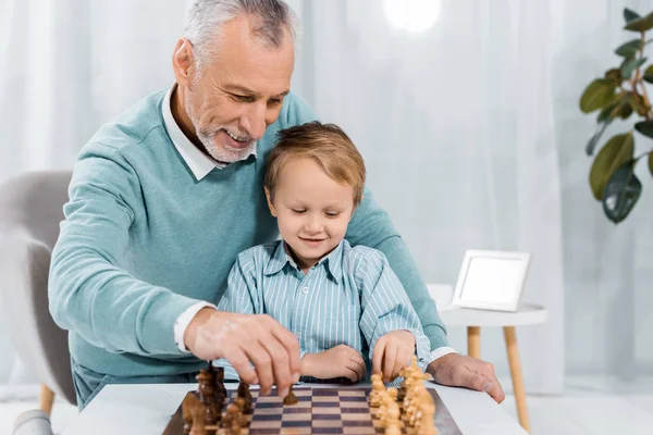 Sonriente hombre de mediana edad enseñando a su nieto a jugar ajedrez en casa - foto de stock