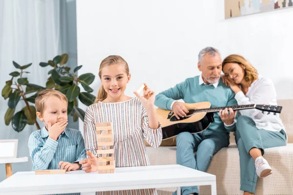 Hermana y hermano jugando bloques de madera torre de juego mientras sus abuelos jugando en la guitarra detrás en casa - foto de stock