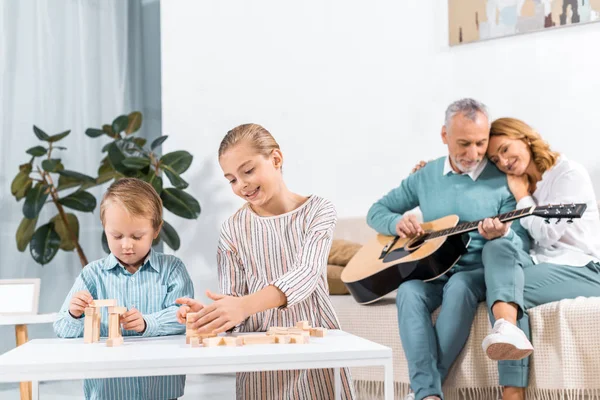 Feliz irmã e irmão jogando blocos de madeira torre de jogo enquanto seus avós tocando na guitarra atrás em casa — Fotografia de Stock