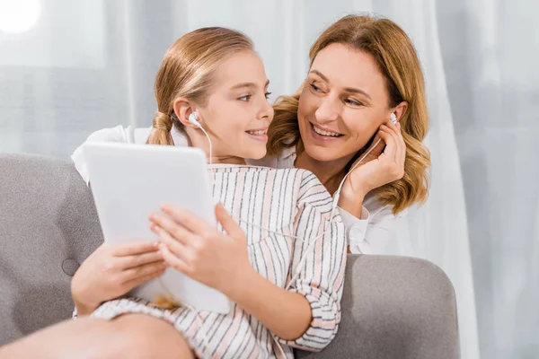 Atractiva mujer sonriente y su nieta en auriculares y tabletas digitales en casa - foto de stock