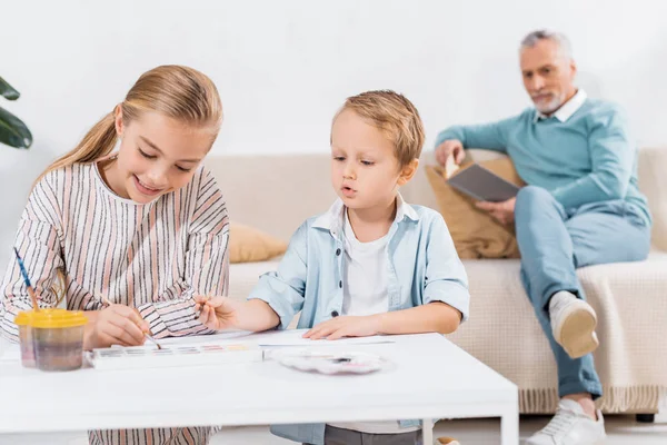 Entzückende Kinder malen am Tisch, während ihr Großvater zu Hause auf dem Sofa ein Buch liest — Stockfoto