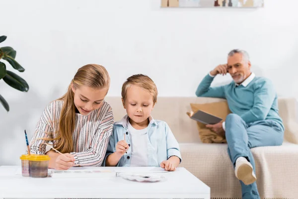 Маленькі діти малюють за столом, а їх дід читає книгу позаду на дивані вдома — стокове фото