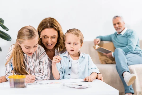 Femme mûre regardant comment ses petits-enfants peignent tandis que son mari assis derrière sur le canapé à la maison — Photo de stock