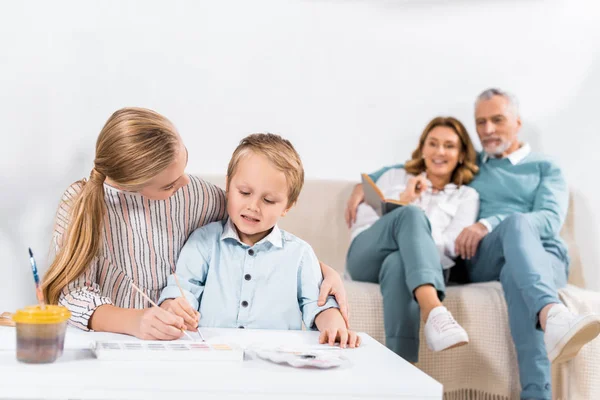 Дитина допомагає маленькому братові малювати за столом, а їхні бабусі і дідусі сидять на дивані позаду вдома — стокове фото