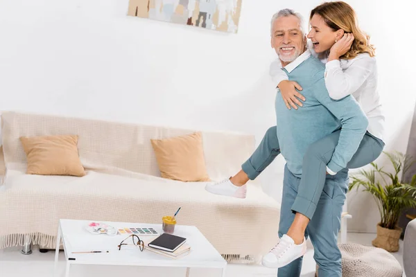 Feliz homem de meia idade fazendo passeio de piggyback para bela esposa na sala de estar em casa — Fotografia de Stock