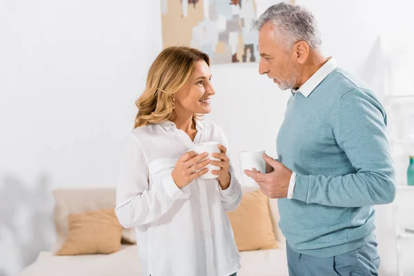 Vista laterale di donna felice con tazza di caffè parlare con marito maturo a casa — Foto stock