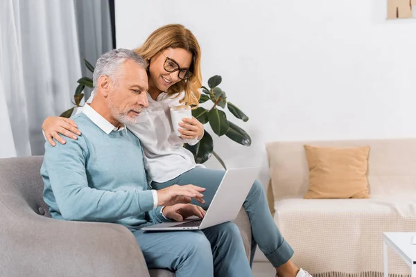 Vista lateral del hombre de mediana edad utilizando el ordenador portátil, mientras que su esposa sonriente sentado cerca con café en casa - foto de stock