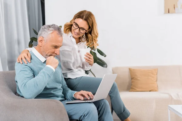 Focused man using laptop while his smiling wife sitting near with coffee at home — Stock Photo