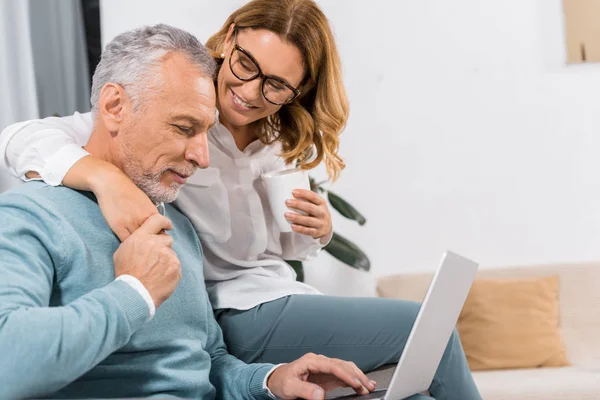 Alegre hombre de mediana edad utilizando el ordenador portátil, mientras que su esposa sonriente sentado cerca con café en casa - foto de stock