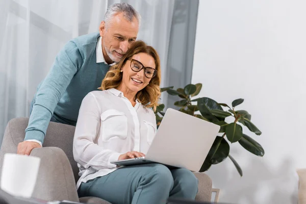 Attractive woman in eyeglasses using laptop while her mature husband standing behind at home — Stock Photo