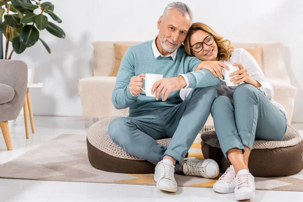 Feliz pareja madura sentada con tazas de café en la sala de estar en casa - foto de stock