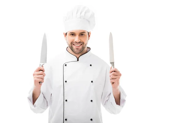 Усміхнений молодий шеф-кухар тримає ріжучі ножі і дивиться на камеру ізольовано на білому — стокове фото
