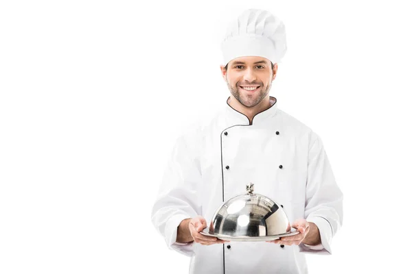 Smiling young chef holding serving tray with dome and looking at camera isolated on white — Stock Photo