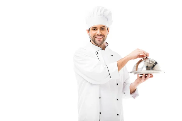 Bonito jovem chef segurando servindo prato com cúpula e olhando para a câmera isolada no branco — Fotografia de Stock