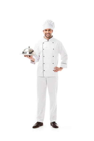 Sorrindo jovem chef segurando prato servindo com cúpula e olhando para a câmera isolada no branco — Fotografia de Stock