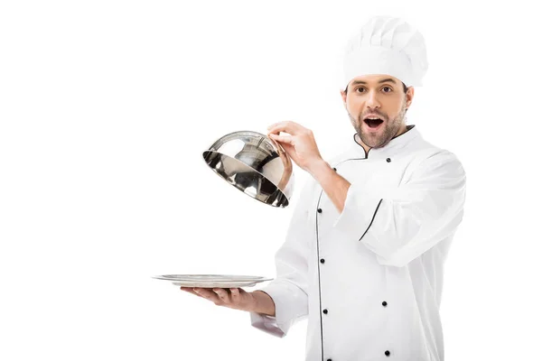 Surprised young chef taking of serving dome from plate and looking at camera isolated on white — Stock Photo