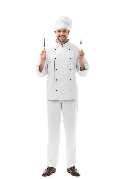 Усміхнений молодий шеф-кухар тримає ніж і виделку і дивиться на камеру ізольовано на білому — стокове фото