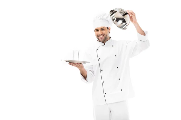 Sonriente joven chef abriendo seving domo con vaso de agua en el interior aislado en blanco - foto de stock