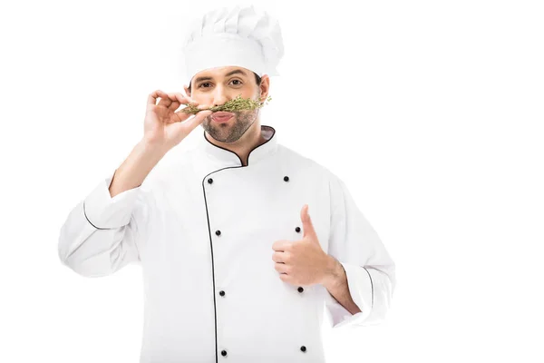 Guapo joven chef haciendo bigote con tomillo y mostrando el pulgar hacia arriba aislado en blanco - foto de stock