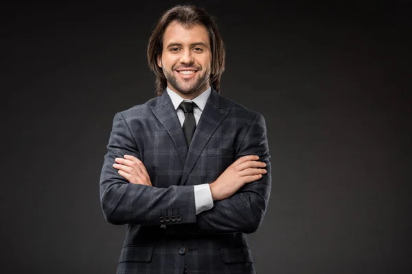 Handsome happy businessman standing with crossed arms and smiling at camera isolated on black — Stock Photo