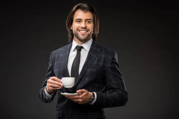 Beau homme d'affaires souriant tenant tasse de café et soucoupe isolé sur noir — Photo de stock