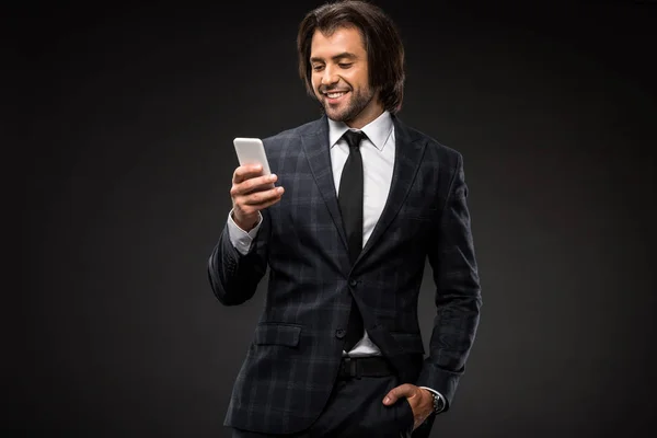 Smiling young businessman standing with hand in pocket and using smartphone isolated on black — Stock Photo