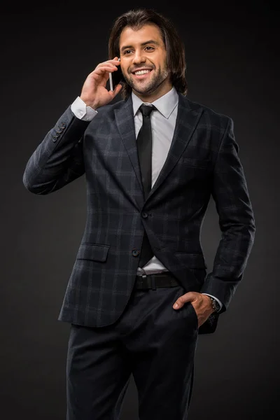 Усміхнений молодий бізнесмен стоїть з рукою в кишені і розмовляє по смартфону ізольовано на чорному — стокове фото