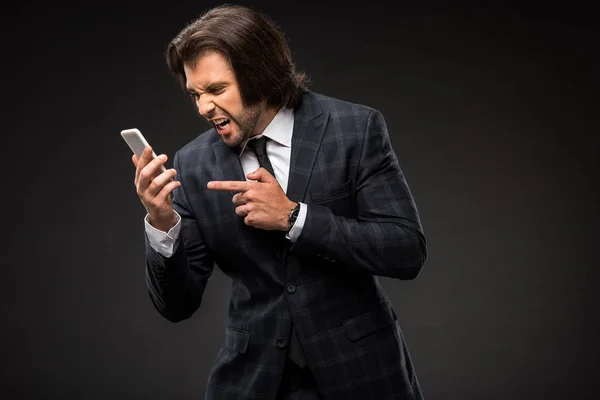 Emotional young businessman screaming at smartphone isolated on black — Stock Photo