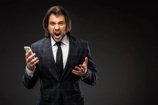 Angry young businessman holding smartphone and yelling at camera isolated on black — Stock Photo