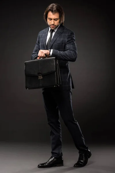 Professional young businessman holding briefcase and checking wristwatch on black — Stock Photo