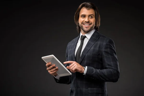 Sonriente joven hombre de negocios usando tableta digital y mirando hacia otro lado aislado en negro - foto de stock