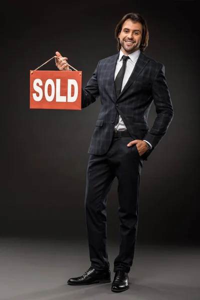 Guapo joven empresario sosteniendo vendido signo y sonriendo a la cámara en negro - foto de stock