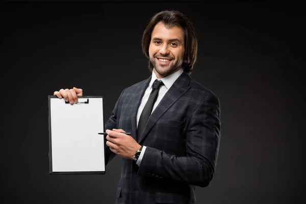 Professional young businessman pointing at blank clipboard and smiling at camera isolated on black — Stock Photo