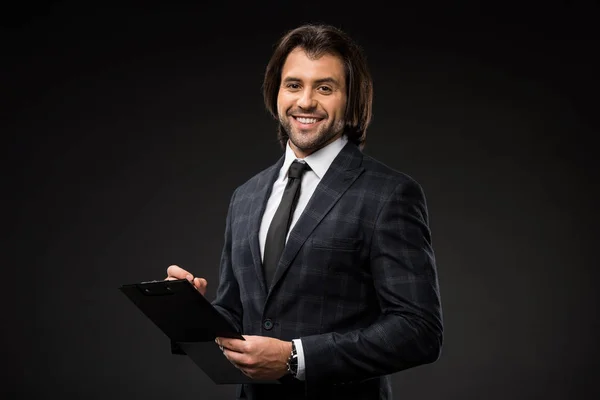 Professional young businessman writing on clipboard and smiling at camera isolated on black — Stock Photo