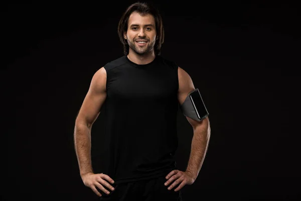 Jeune homme athlétique avec brassard et smartphone debout avec les mains sur la taille et souriant à la caméra isolée sur noir — Photo de stock