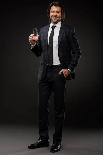 Vista completa de joven hombre de negocios guapo sosteniendo un vaso de agua y sonriendo a la cámara en negro - foto de stock