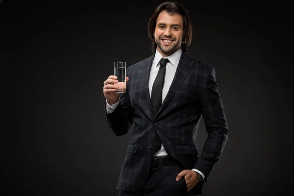 Joven hombre de negocios guapo sosteniendo un vaso de agua y sonriendo a la cámara aislado en negro - foto de stock