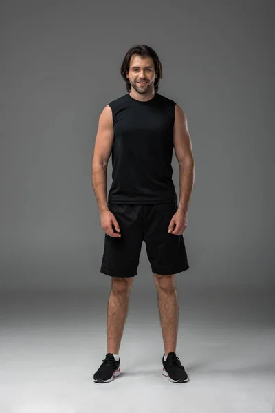 Full length view of handsome muscular young sportsman standing and smiling at camera on grey — Stock Photo