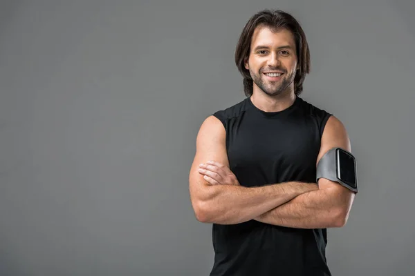 Jeune homme athlétique avec brassard et smartphone debout avec bras croisés et souriant à la caméra isolée sur gris — Photo de stock