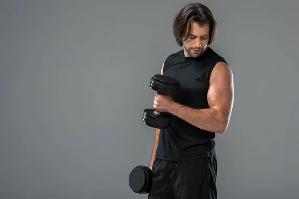 М'язистий красивий чоловік тренується з гантелями і дивиться на біцепси ізольовані на сірому — стокове фото