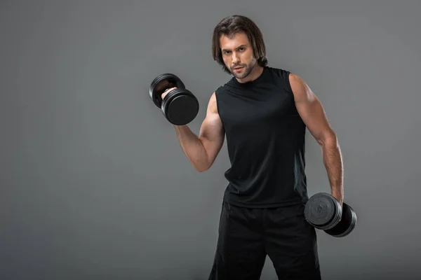 Athletic young sportsman exercising with dumbbells and looking at camera isolated on grey — Stock Photo