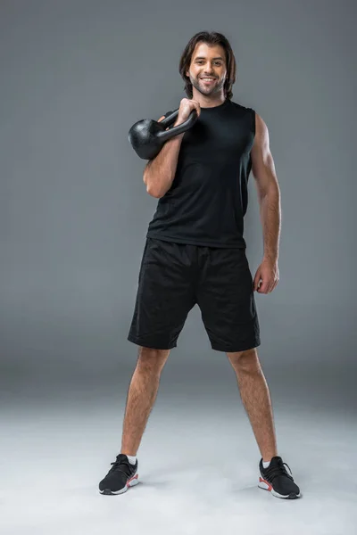 Ganzkörperansicht des athletischen jungen Mannes, der mit der Kettlebell trainiert und auf grau in die Kamera lächelt — Stockfoto