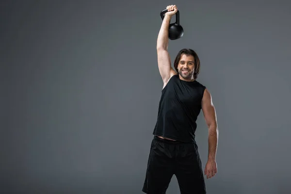 Muskulöser junger Mann trainiert mit Kettlebell und lächelt in die Kamera isoliert auf grau — Stockfoto