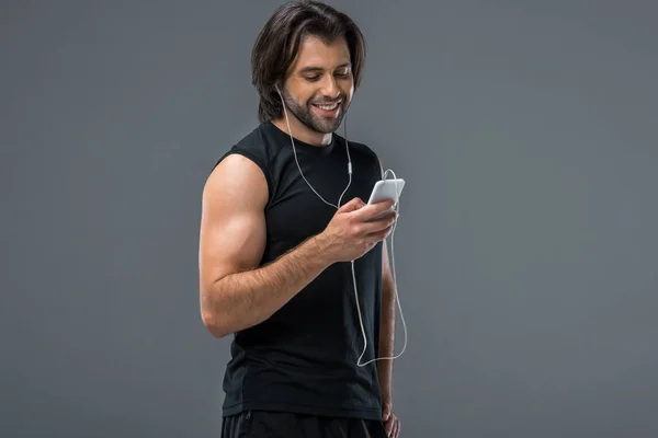 Uomo muscoloso sorridente in auricolare utilizzando smartphone isolato su grigio — Foto stock