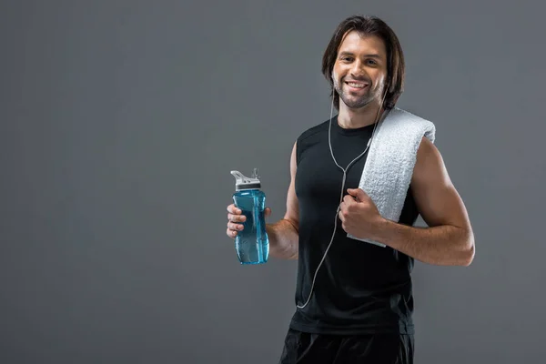 Счастливый молодой спортсмен в наушниках, держа бутылку воды и улыбаясь в камеру, изолированную на сером — стоковое фото